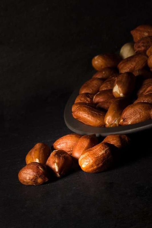 Raw peanuts in their brown outer shells, with some scattered on a dark surface and others piled on a dark plate against a black background.