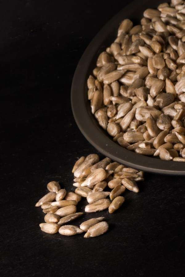 Raw sunflower seeds spilled from a dark ceramic plate onto a dark black surface, with more seeds visible inside the plate.